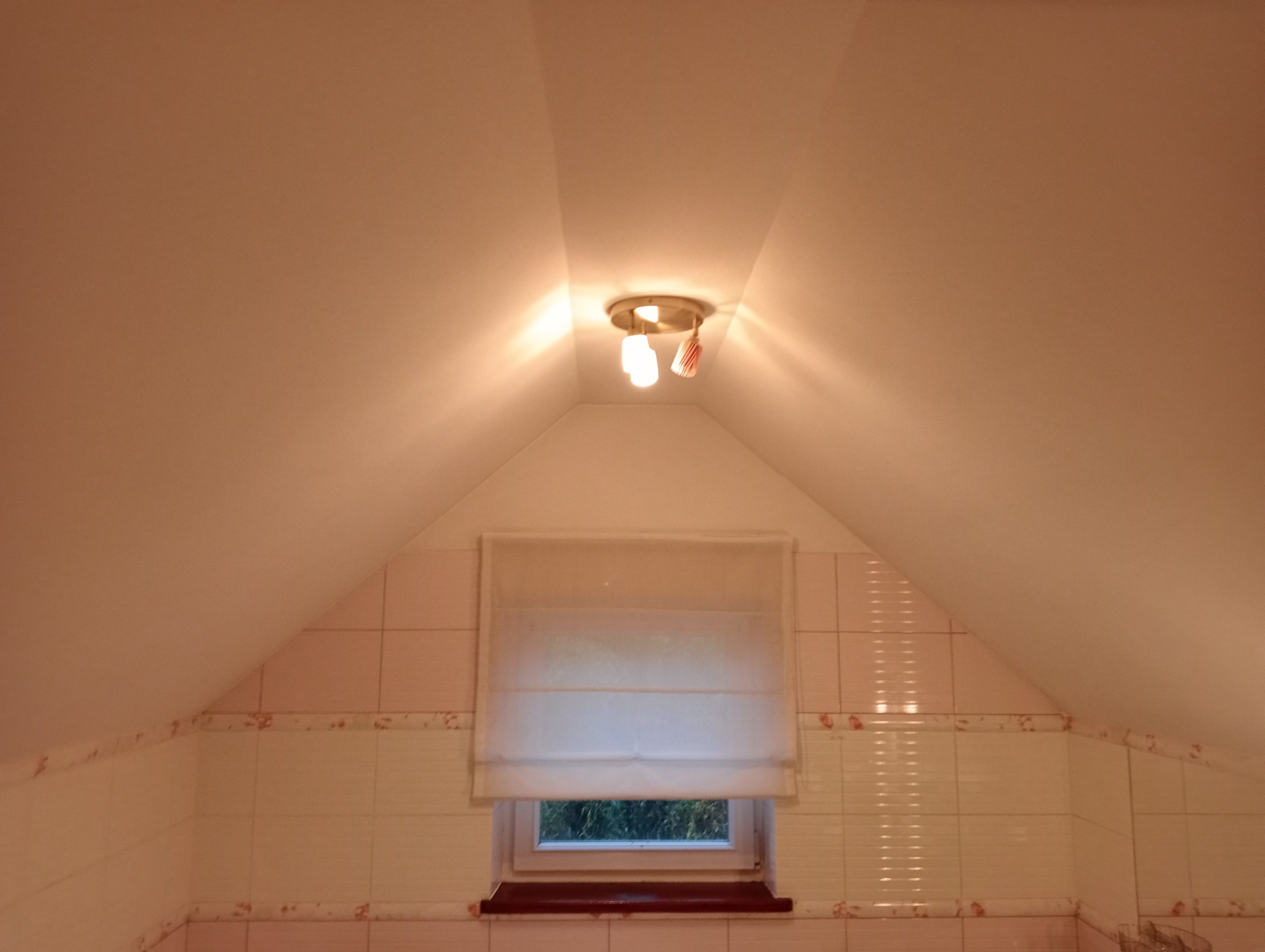 Ukośna ściana poddasza z oknem zasłoniętym roletą rzymską i oświetleniem sufitowym z trzema żarówkami