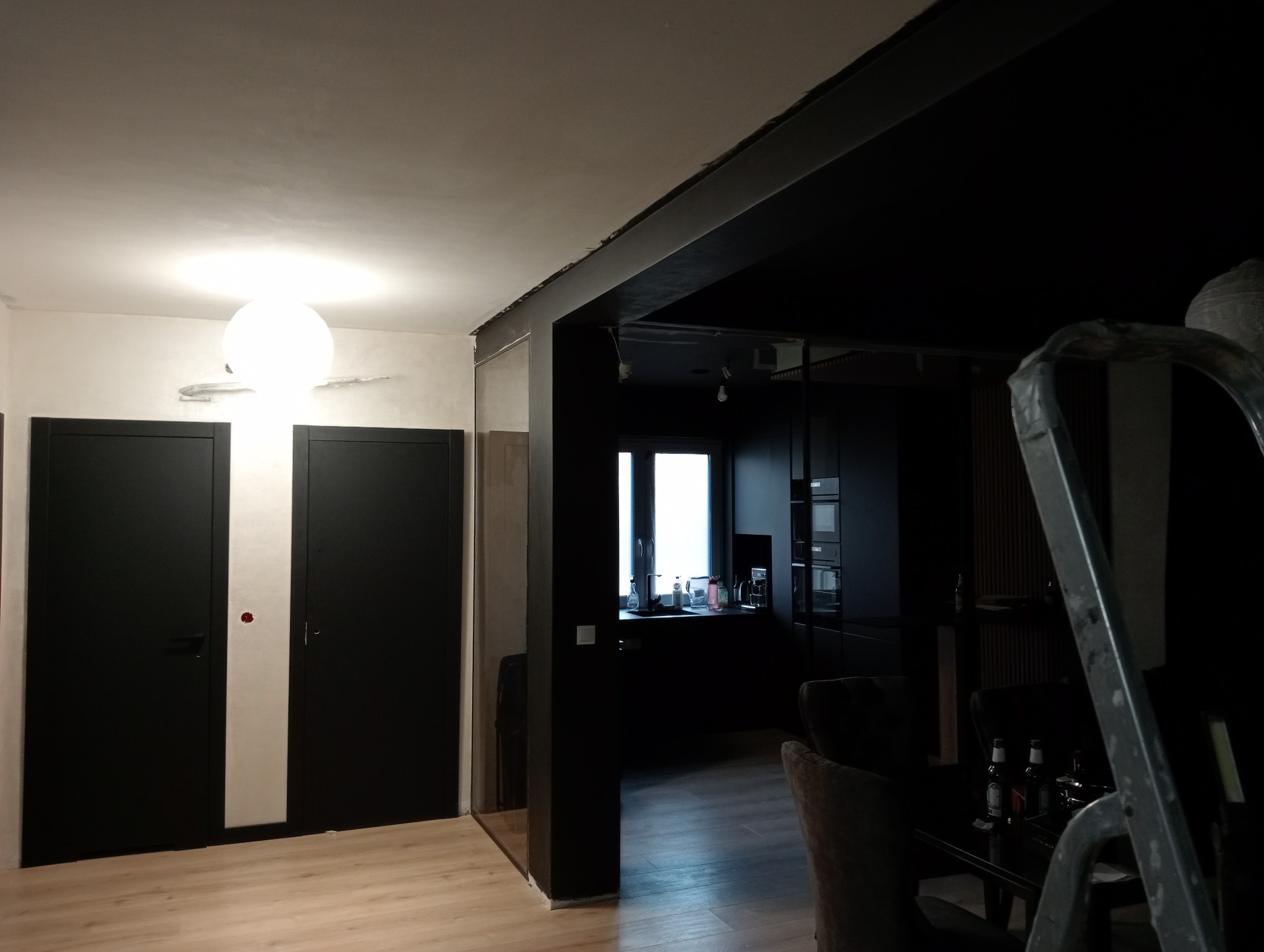 Wnętrze mieszkania w trakcie prac wykończeniowych: widoczne gładzie na suficie, czarne drzwi i fragment kuchni w ciemnej tonacji, zarys drabiny malarskiej.