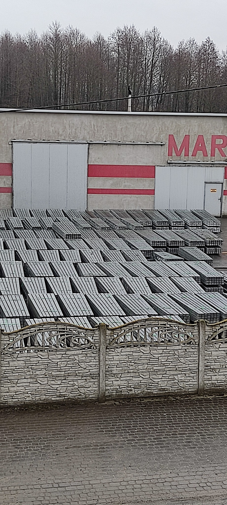 Stosy prefabrykowanych paneli ogrodzeniowych z betonu architektonicznego składowane obok budynku magazynowego z czerwonymi pasami.