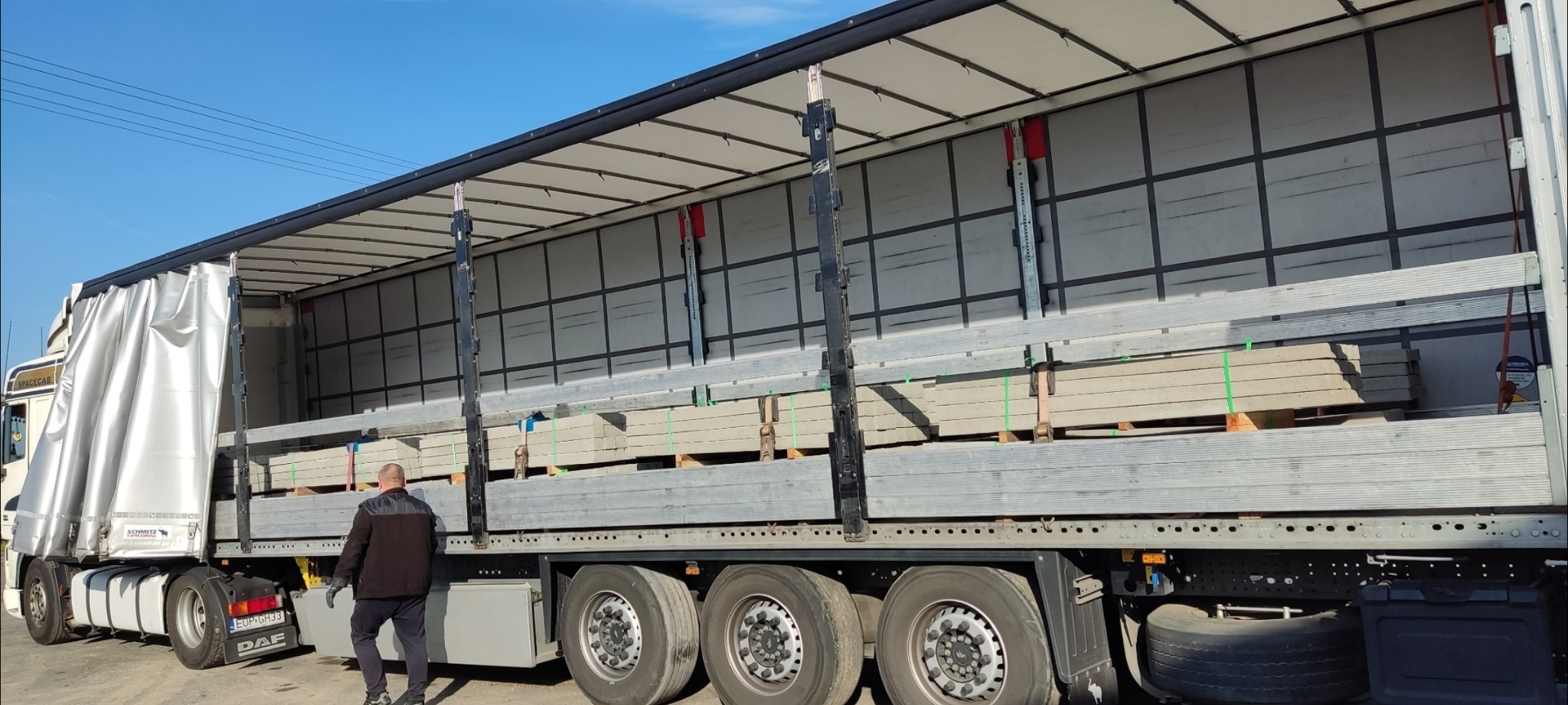 Otwarta naczepa ciężarówki z załadunkiem palet z betonowymi elementami ogrodzeniowymi, zabezpieczonymi taśmami, widoczny pracownik w czarnej kurtce i rękawicach.