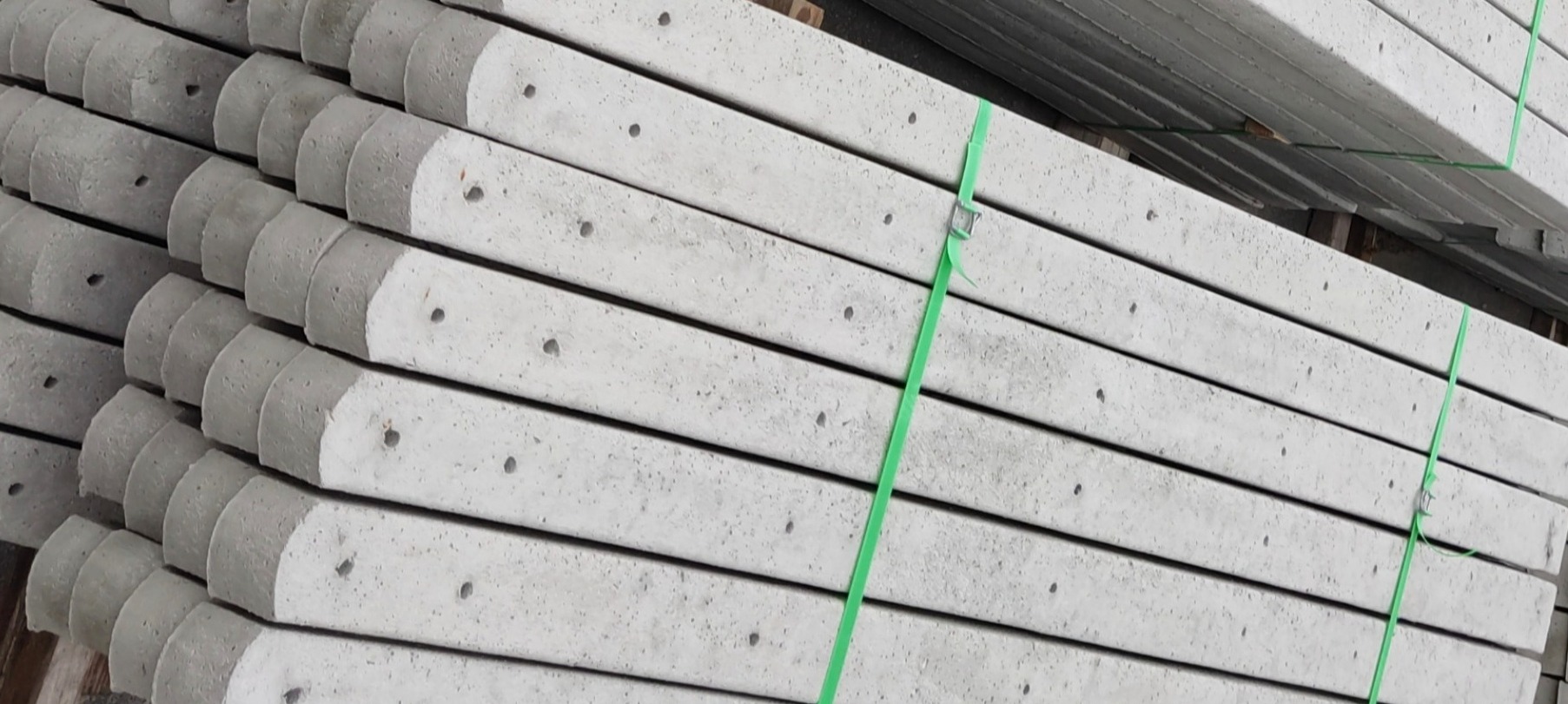 Stos betonowych słupków ogrodzeniowych z zaokrąglonymi krawędziami, spiętych zieloną taśmą, widoczne otwory montażowe.