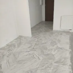 Jasne, marmurowe płytki podłogowe ułożone w korytarzu z białymi ścianami i brązowymi drzwiami wejściowymi, widoczny grzejnik.