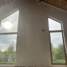 Wnętrze domu w trakcie prac wykończeniowych, ściany po nałożeniu gładzi szpachlowej, widoczne dwa okna o nietypowym kształcie i drewnianych ramach, przez które widać zieleń ogrodu.