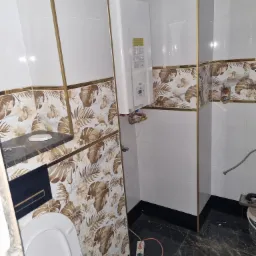 Wnętrze łazienki w trakcie prac wykończeniowych: biało-złote płytki z motywem liści, czarna marmurowa podłoga, biały bojler na ścianie, widoczne przewody i narzędzia glazurnicze.