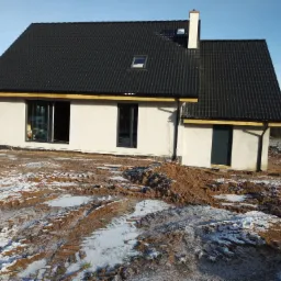 Nowo wybudowany dom z czarną dachówką, jasną elewacją i białym kominem, otoczony ziemią z resztkami śniegu, widoczne ślady opon.