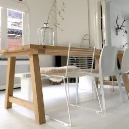 Jasne wnętrze jadalni w Łomiankach: drewniany stół na kozłach, białe krzesła o ażurowej konstrukcji, biała podłoga, za stołem kominek i dekoracje ścienne, w tym poroże jelenia.