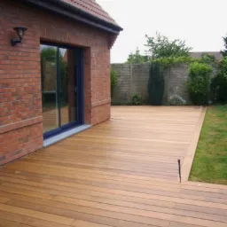 Jasnobrązowy drewniany taras przylegający do ceglanego domu z niebieskimi drzwiami przesuwnymi, otoczony zielonym trawnikiem i betonowym murem z roślinnością.