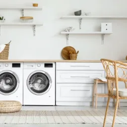 Jasne pomieszczenie gospodarcze z dwiema pralkami pod blatem, białymi szafkami, wiklinowym pufem i krzesłem, oraz półkami z dekoracjami.