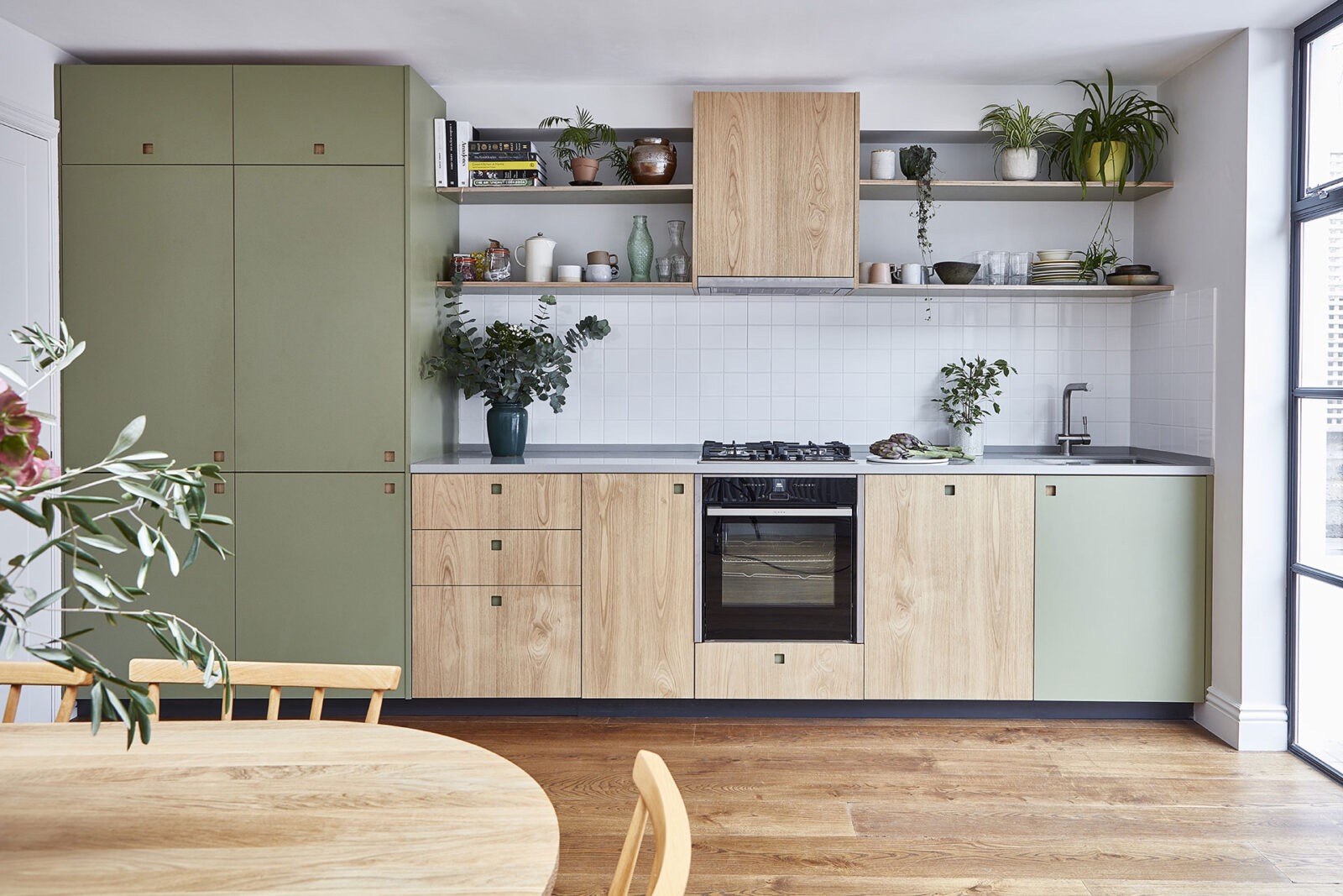 Nowoczesna kuchnia z drewnianymi frontami szafek i zielonymi akcentami, uzupełniona o drewniany stół i półki z roślinami.