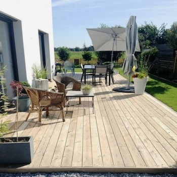 Drewniany taras z jasnych desek, otoczony białym domem i zielonym trawnikiem. Na tarasie wiklinowe fotele, stół z krzesłami pod dużym parasolem, dekoracyjne donice z roślinami.