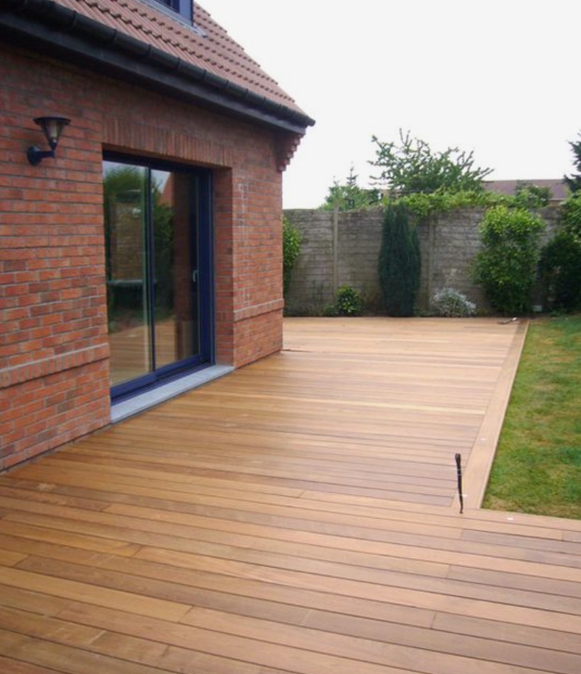 Jasnobrązowy drewniany taras przylegający do ceglanego domu z niebieskimi drzwiami przesuwnymi, otoczony zielonym trawnikiem i betonowym murem z roślinnością.