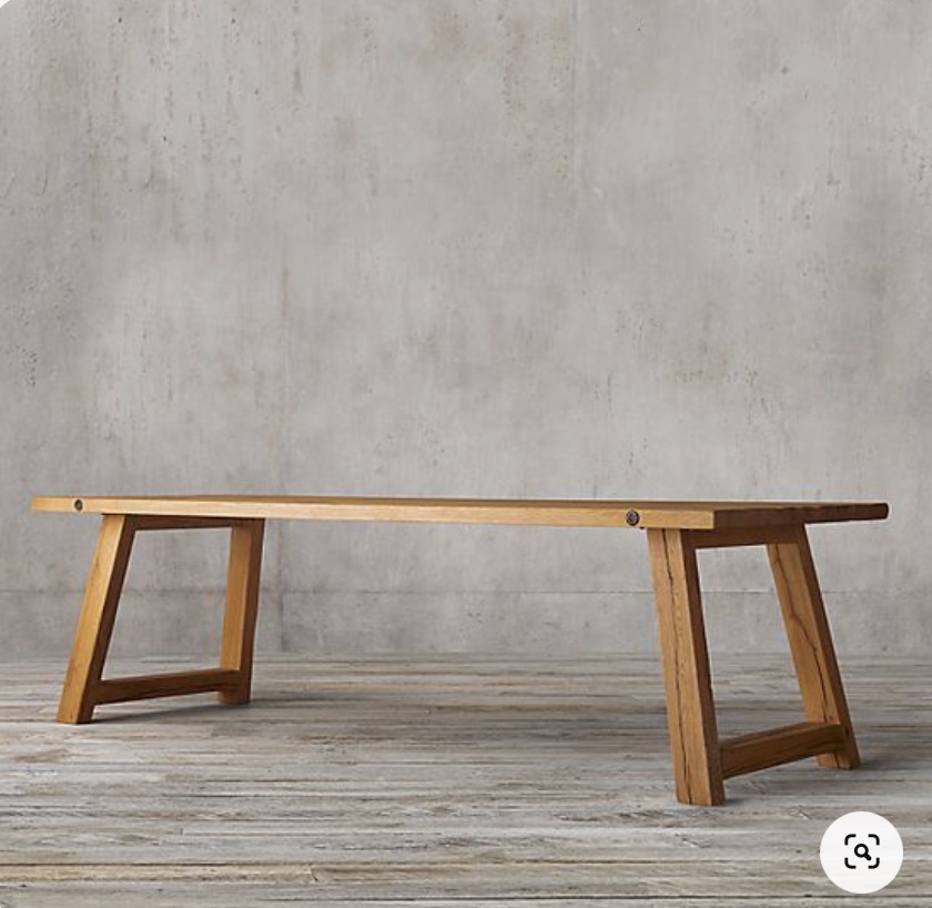 Prosty, drewniany stół z jasnego drewna, z widocznymi śrubami mocującymi nogi, ustawiony na podłodze z desek na tle szarej, teksturowanej ściany.