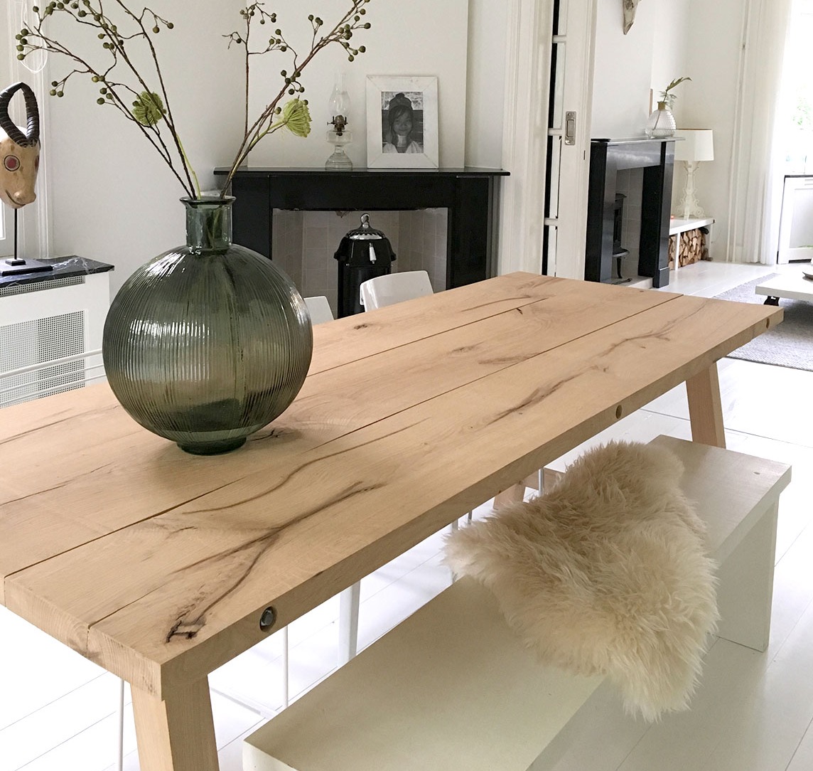 Jasny drewniany stół jadalny z widocznym usłojeniem, ustawiony na tle białego wnętrza z kominkiem i dekoracjami, z zielonym wazonem z gałązkami na blacie i futrzaną poduszką na ławce.