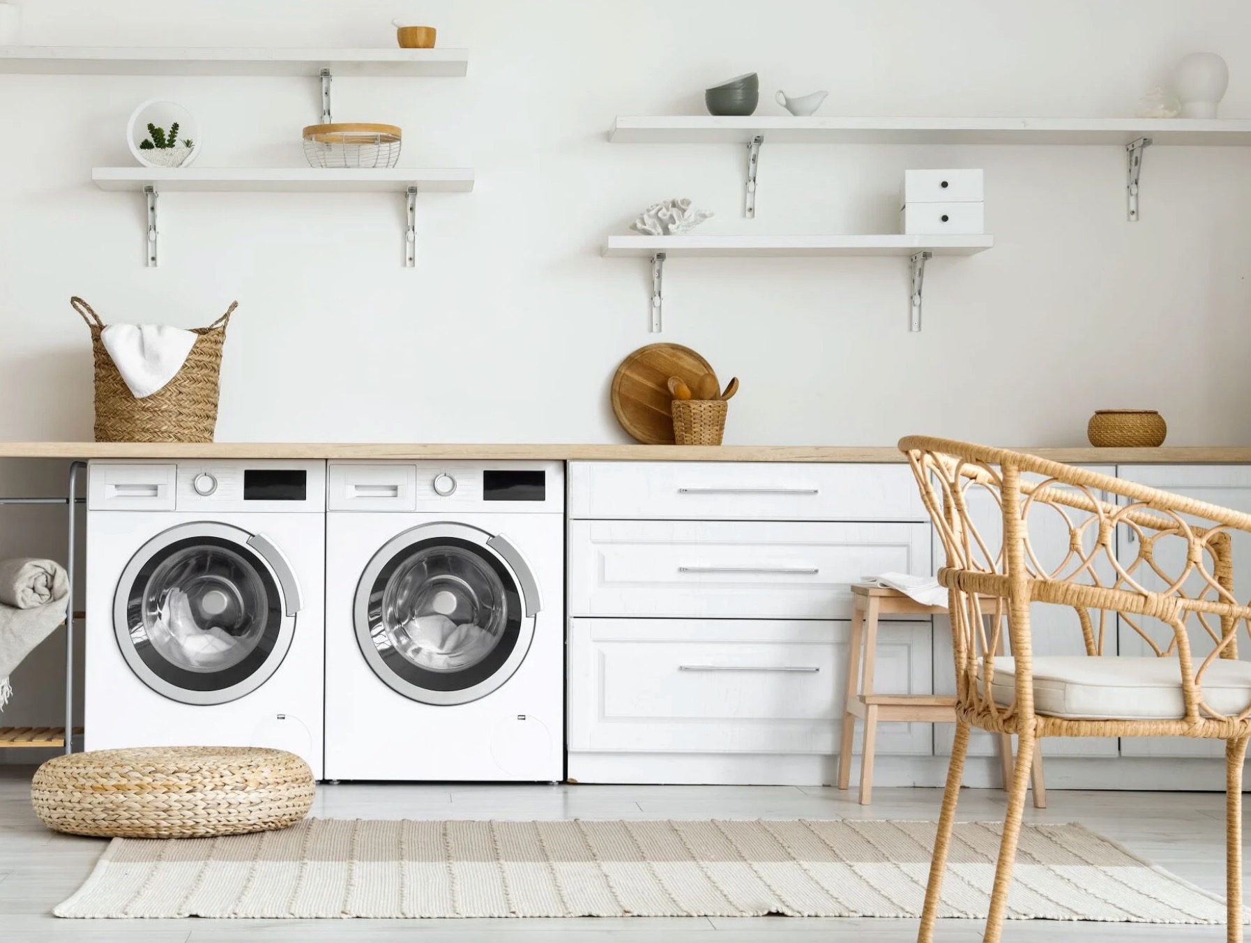 Jasne pomieszczenie gospodarcze z dwiema pralkami pod blatem, białymi szafkami, wiklinowym pufem i krzesłem, oraz półkami z dekoracjami.