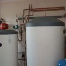 Dwa białe, cylindryczne zbiorniki ciepłej wody użytkowej z szarymi pokrywami, połączone miedzianymi rurami i zaworami, zamontowane w pomieszczeniu technicznym z jasnymi ścianami. Widoczne także...