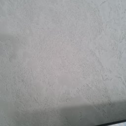 KN-BUD Krzysztof Nyczka - Szara ściana z wyraźną, nieregularną fakturą, imitującą efekt dekoracyjny, z fragmentem telewizora w dolnym rogu kadru.