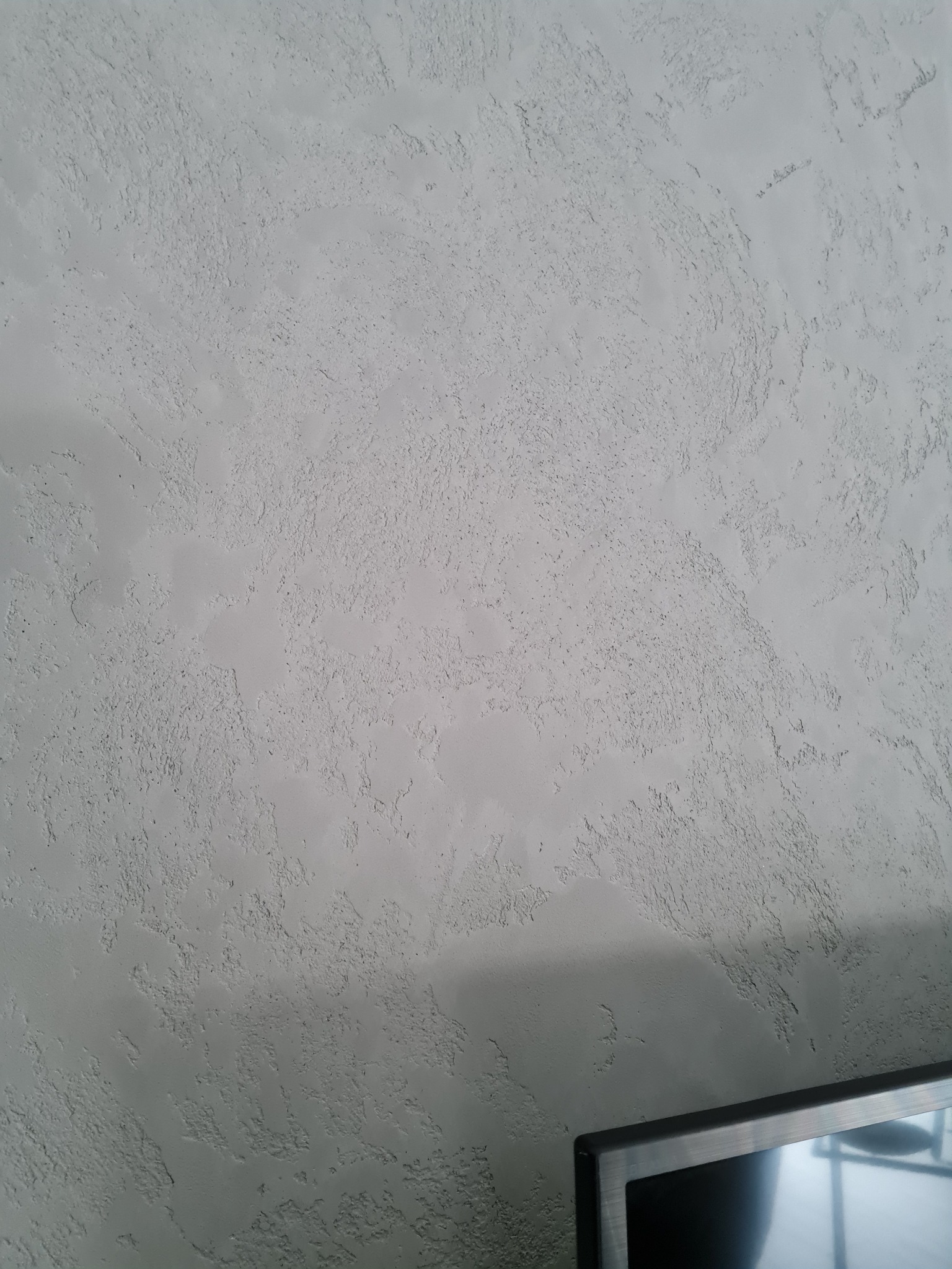 Szara ściana z wyraźną, nieregularną fakturą, imitującą efekt dekoracyjny, z fragmentem telewizora w dolnym rogu kadru.