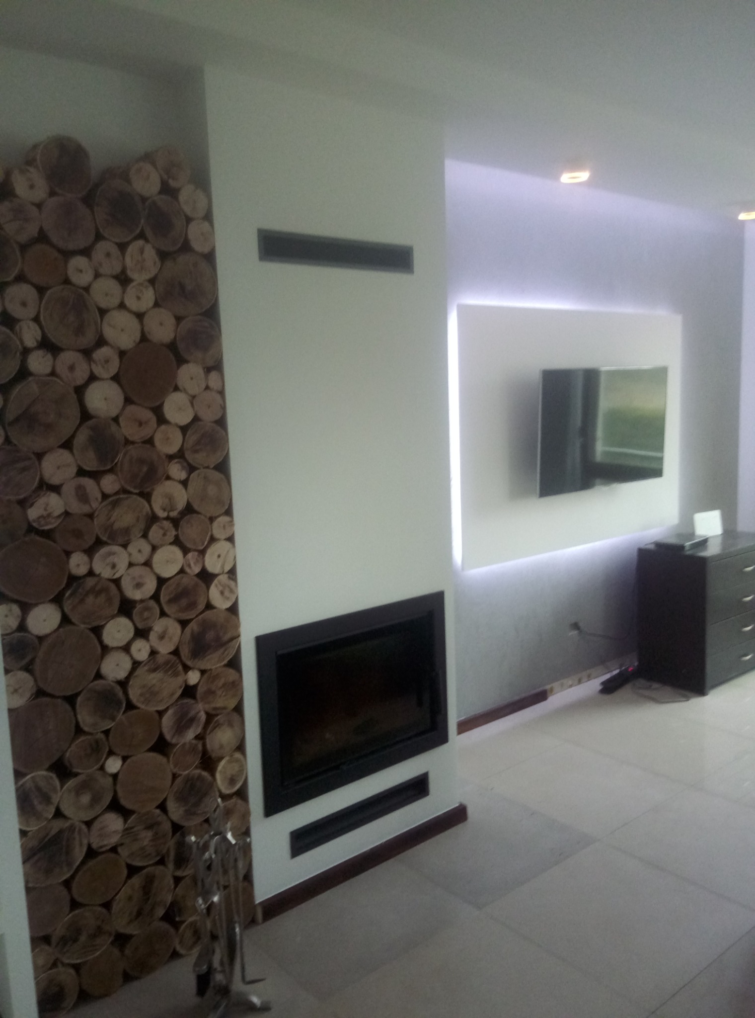 Nowoczesny salon z kominkiem w czarnej obudowie, ściana wykończona dekoracyjnymi plastrami drewna, podświetlony telewizor i biała podłoga z płytek.