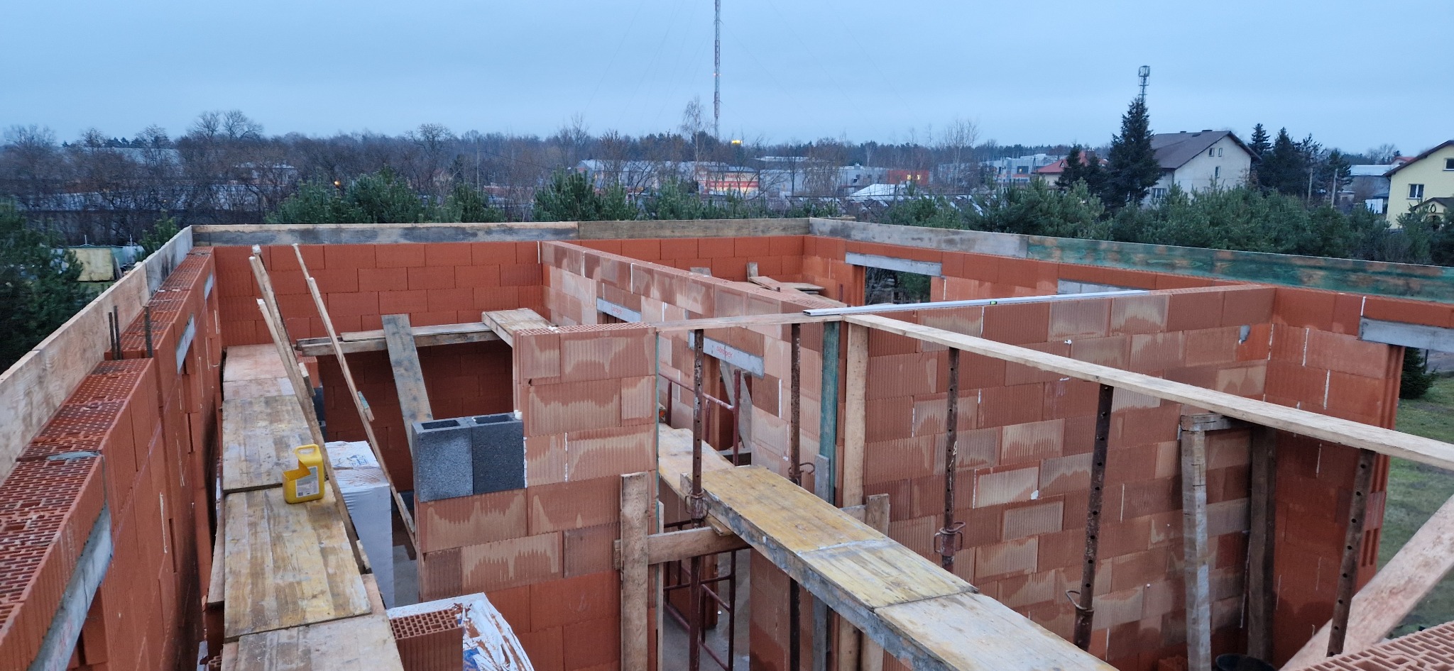 Wnętrze budowanego domu z czerwonej cegły ceramicznej; widoczne drewniane rusztowania i podpory; w tle drzewa i zabudowania.