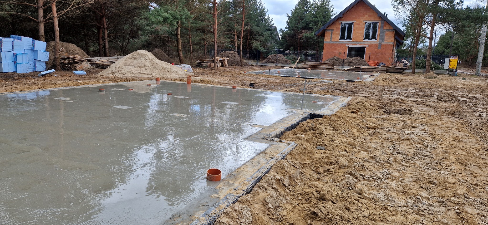 Wylany fundament pod budynek murowany z widocznymi instalacjami i izolacją, w tle częściowo wzniesione ściany z czerwonej cegły i stosy materiałów budowlanych.