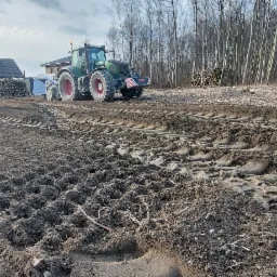 Przygotowanie ziemi pod trawnik: ślady opon traktora na świeżo przekopanej ziemi, w tle dom i sterta pociętych drzew