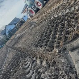 Przygotowane pole pod trawnik z rolki, widoczne ślady opon traktora, w tle domy i drzewa bez liści, pochmurne niebo.