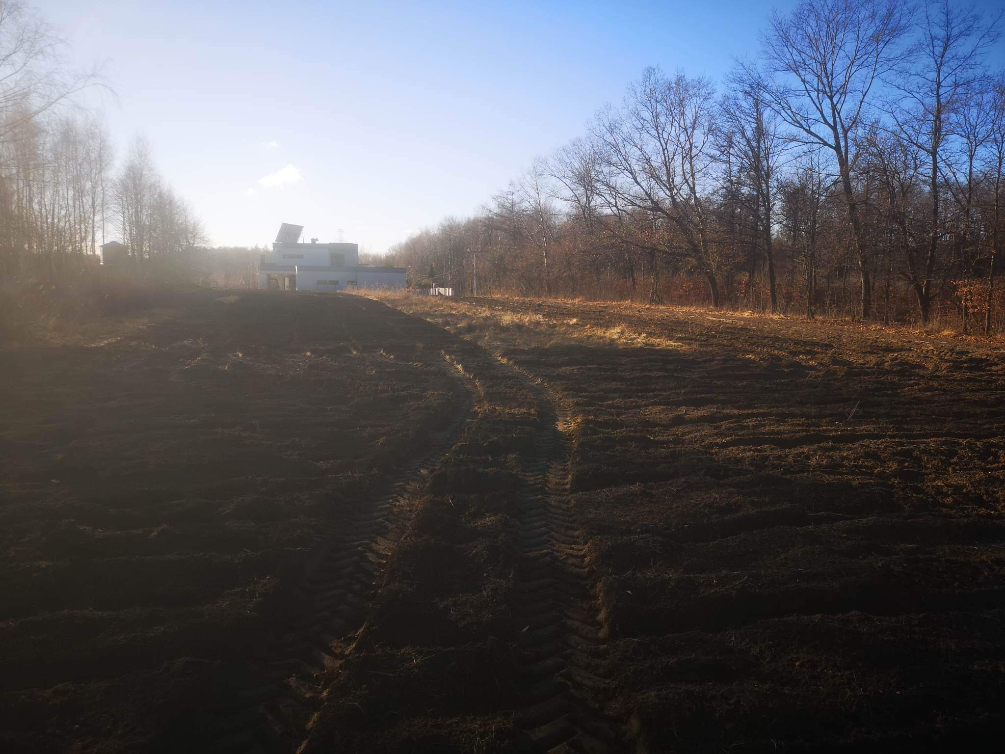 Świeżo przekopana, ciemna ziemia z wyraźnymi śladami opon ciągnika, przygotowana pod przyszły trawnik lub uprawę, budynek w tle w oddali.