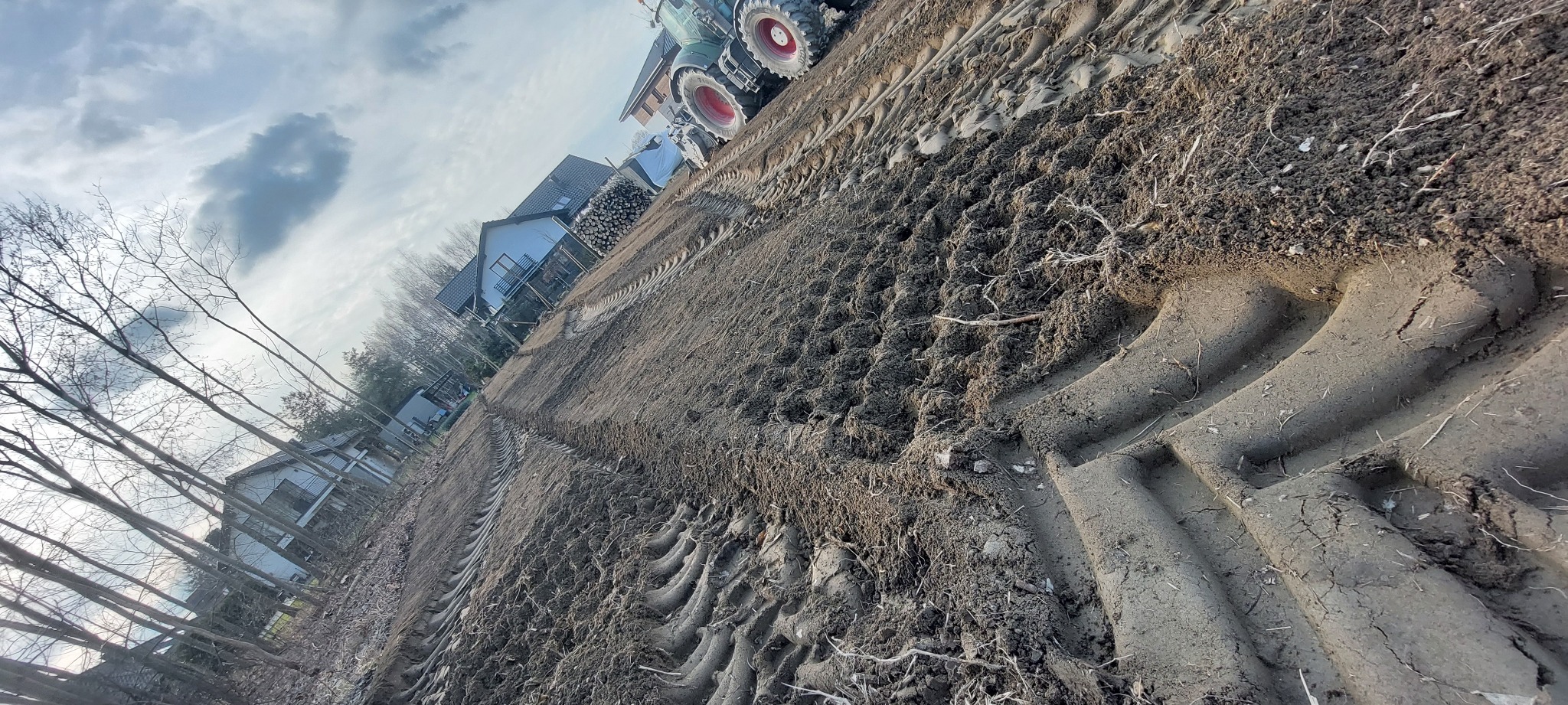 Przygotowane pole pod trawnik z rolki, widoczne ślady opon traktora, w tle domy i drzewa bez liści, pochmurne niebo.