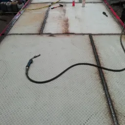 Fragment metalowej platformy z blachy ryflowanej, spawana wzdłuż łączeń, widoczne narzędzia i akcesoria spawalnicze, w tle pojemniki z olejami.