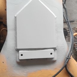 S Technik Sergiy Naumov - Fragment pomalowanej na szaro metalowej konstrukcji maszyny budowlanej z widocznymi spawami i mocowaniami, z butem osoby stojącej obok