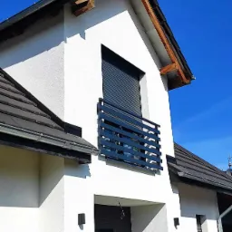 Nowoczesna balustrada balkonowa w kolorze grafitowym, z poziomych profili, zamontowana na białym budynku z ciemną dachówką, widok z dołu na błękitne niebo.