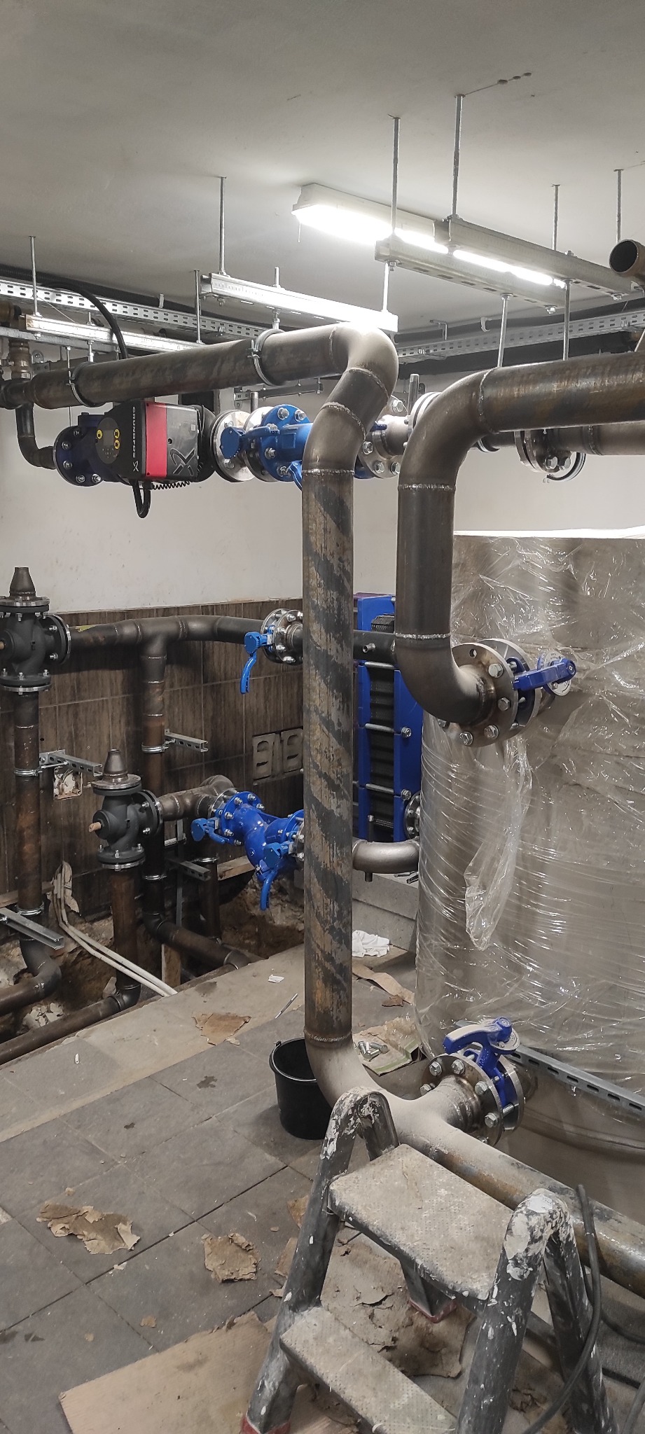 Instalacja grzewcza w kotłowni z rurami, zaworami, pompą obiegową i wymiennikiem ciepła, widoczna drabina malarska i ślady po remoncie na podłodze.