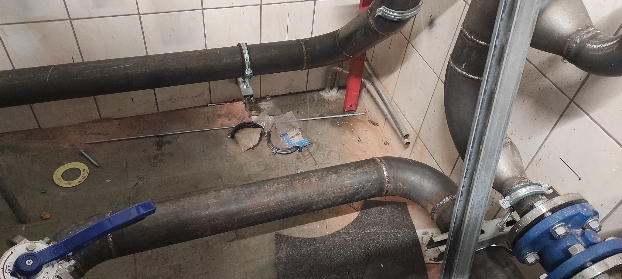 Surowe, metalowe rury połączone spawami, z widocznymi elementami mocującymi i zaworem z niebieską rączką, w trakcie instalacji w pomieszczeniu wyłożonym kafelkami.