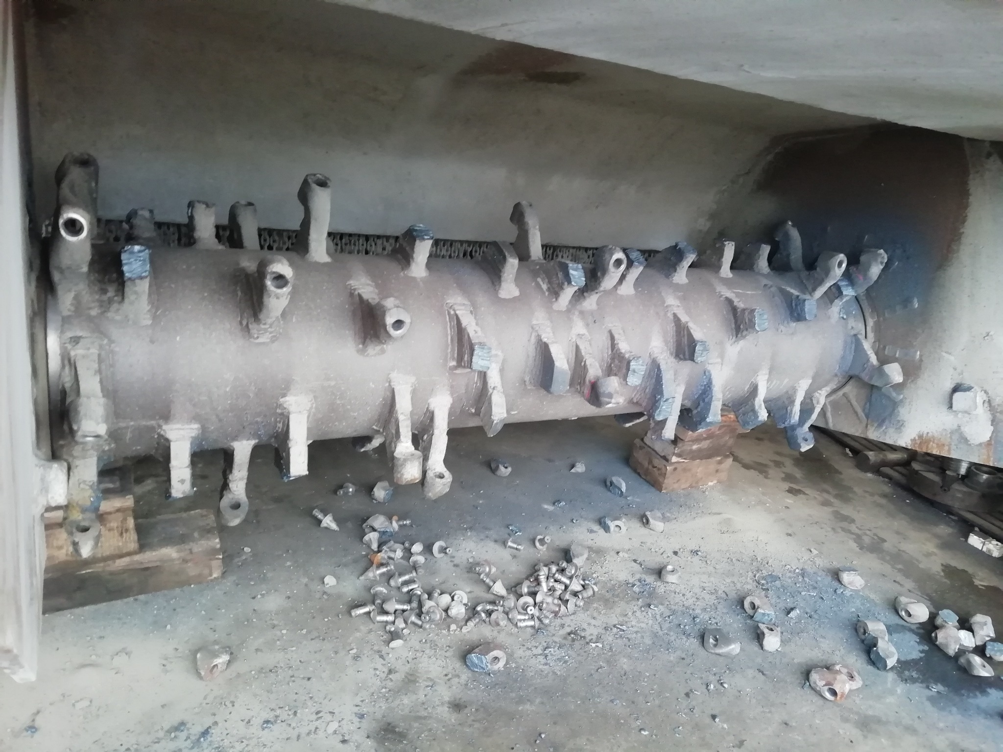 Cylindryczny, metalowy element roboczy z licznymi uchwytami, prawdopodobnie fragment większej maszyny, leżący na drewnianych klockach, z rozrzuconymi śrubami i nakrętkami na betonowej posadzce.