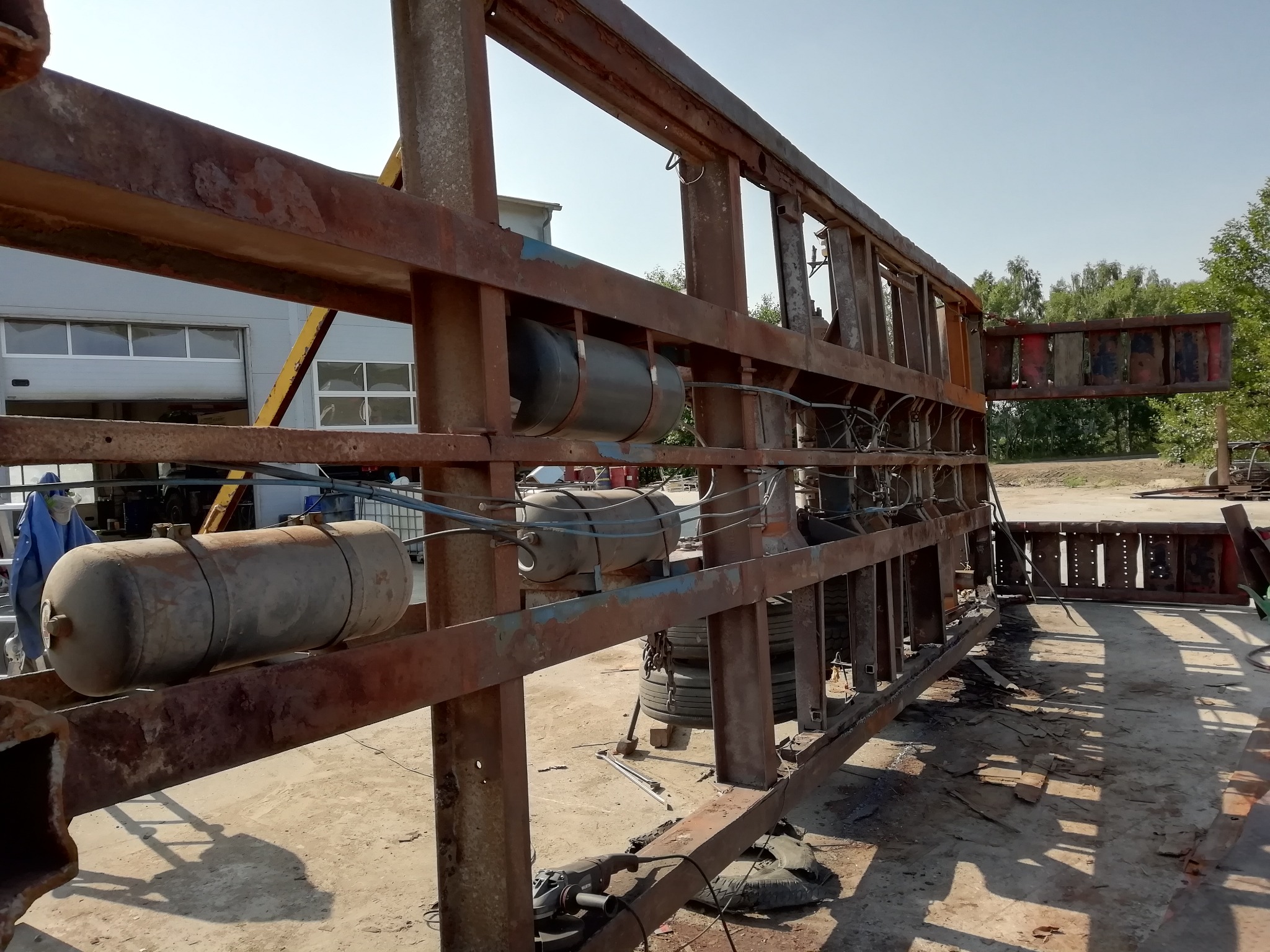 Konstrukcja metalowa z elementami zbiorników, widoczne ślady rdzy i spawania, w tle budynek warsztatowy i zieleń.