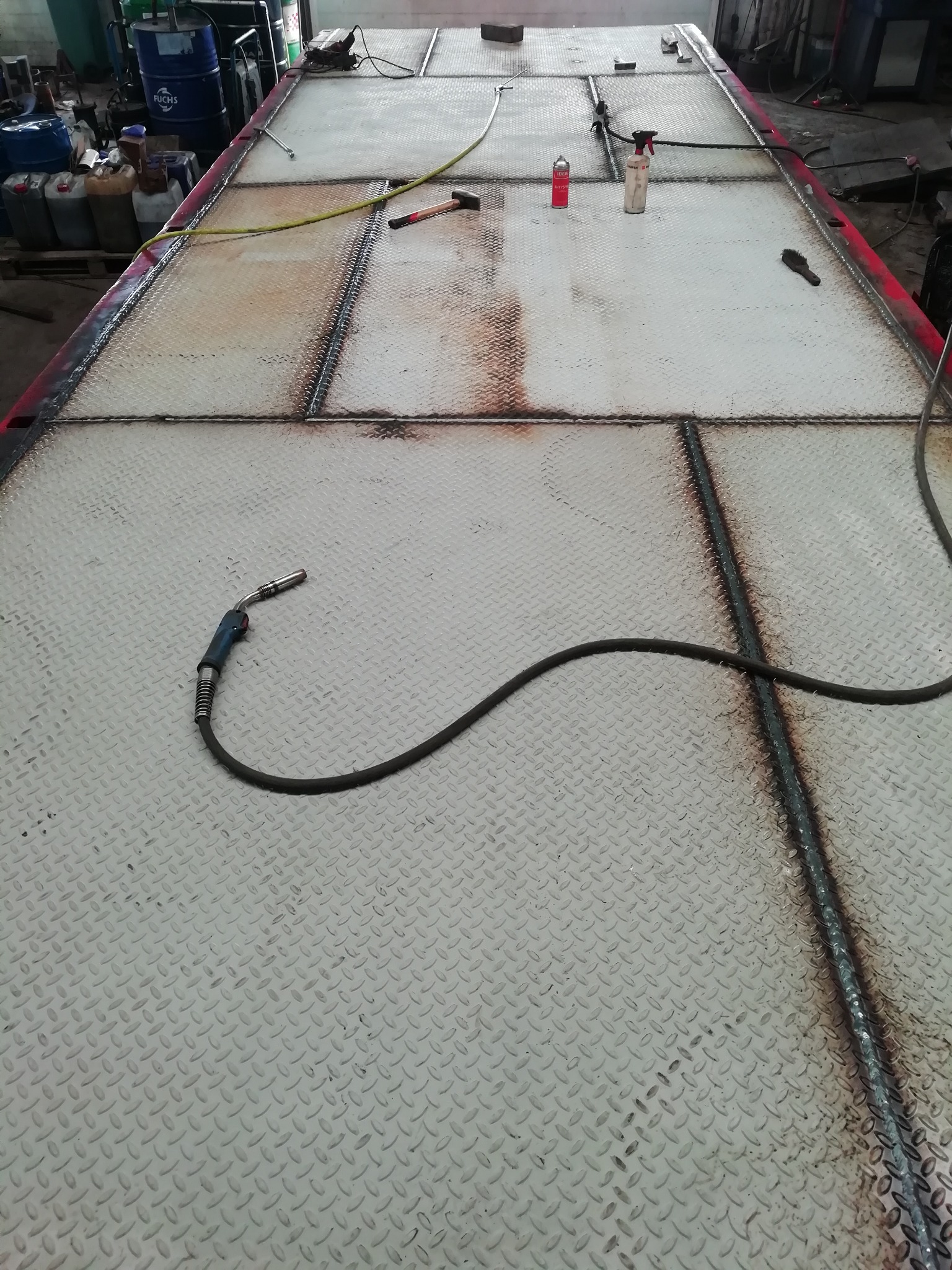 Fragment metalowej platformy z blachy ryflowanej, spawana wzdłuż łączeń, widoczne narzędzia i akcesoria spawalnicze, w tle pojemniki z olejami.