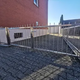 Otwarta, dwuskrzydłowa brama wjazdowa ze stali, z pionowymi prętami zakończonymi kulkami, na posesji z kostki brukowej, widok z zewnątrz.