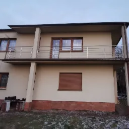 Dwupiętrowy dom z beżową elewacją, z balkonem zabezpieczonym poziomą, metalową konstrukcją, widok z zewnątrz w pochmurny dzień.