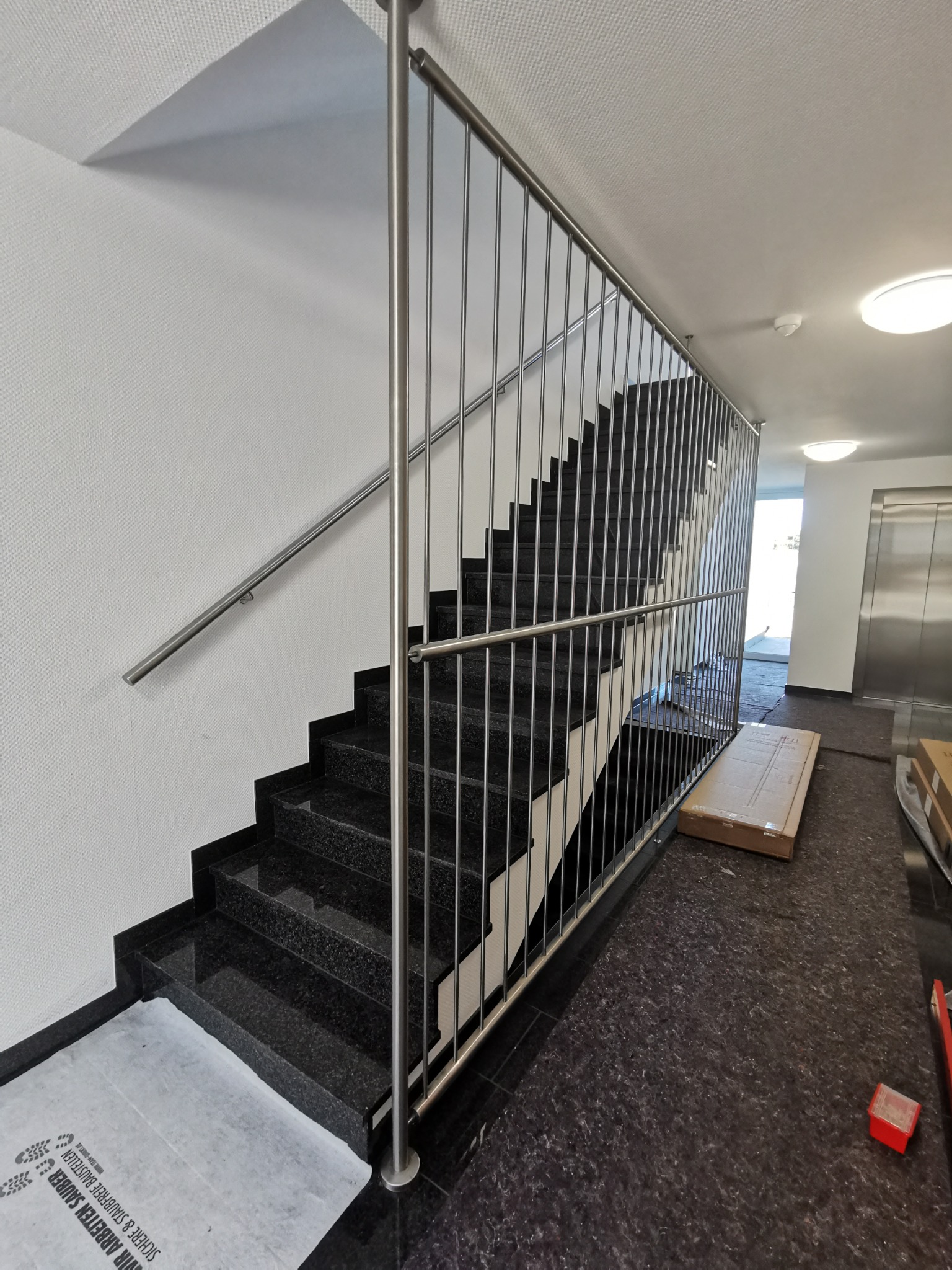 Schody z czarnego granitu z pionową, metalową barierką ochronną i poręczą, zabezpieczone folią ochronną na podłodze i schodach, w tle winda.