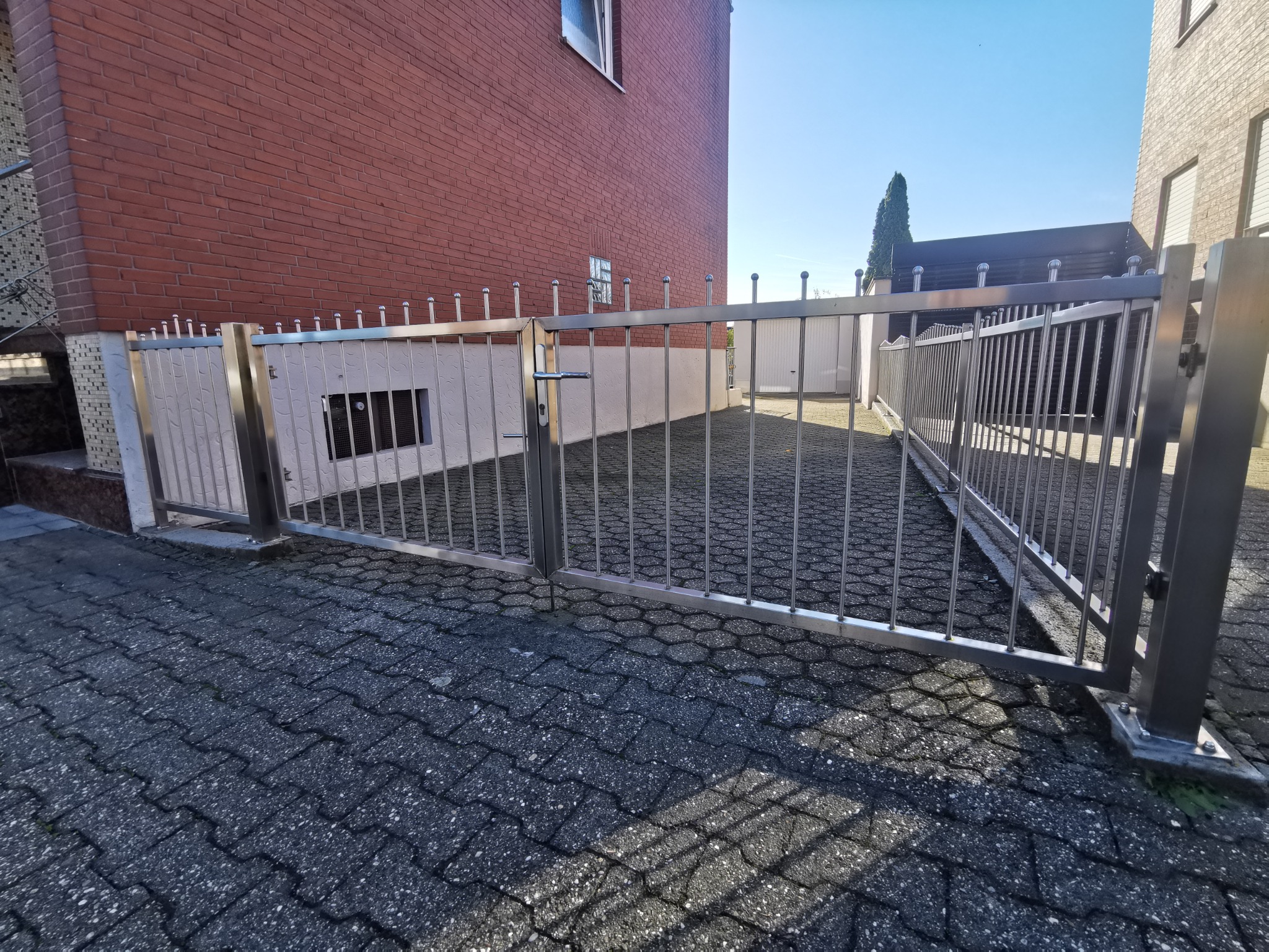 Otwarta, dwuskrzydłowa brama wjazdowa ze stali, z pionowymi prętami zakończonymi kulkami, na posesji z kostki brukowej, widok z zewnątrz.