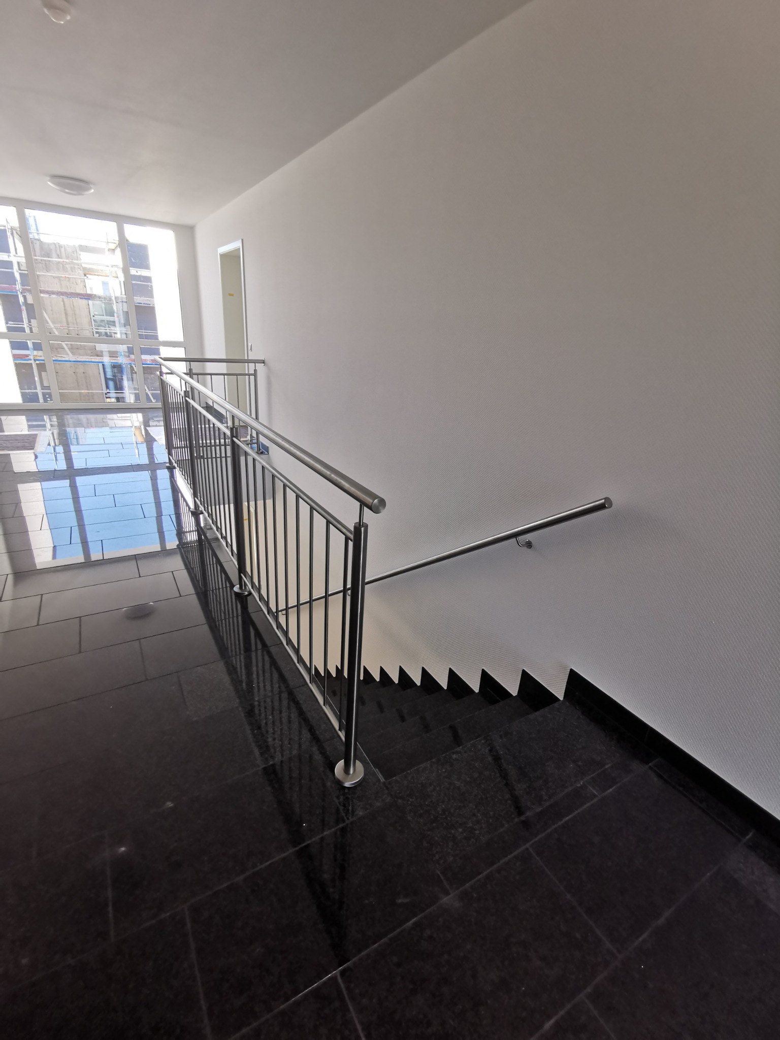 Schody wyłożone czarnym granitem z metalową balustradą i poręczą, widoczne okno z rusztowaniami na zewnątrz.