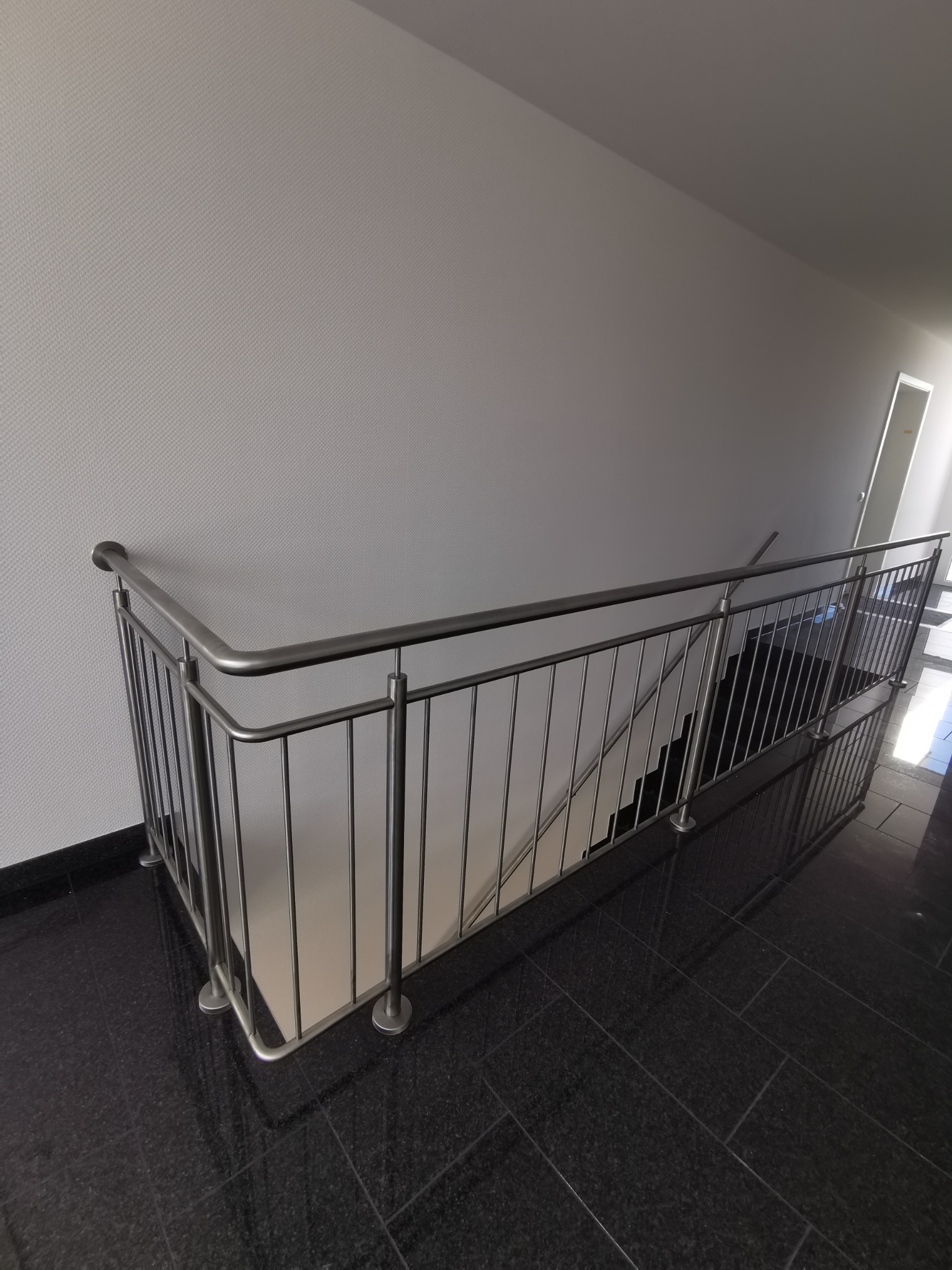Szczegół ujęcia schodów z metalową balustradą o minimalistycznym designie, z polerowaną czarną podłogą z płytek i jasną ścianą w tle.