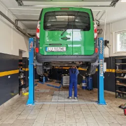 Zielony samochód dostawczy na podnośniku dwukolumnowym w warsztacie, widok z tyłu na tablicę rejestracyjną i pracownika w kombinezonie.