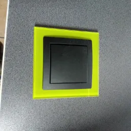 Czarny włącznik światła z podświetlaną, neonowo-zieloną ramką na szarej, teksturowanej ścianie.