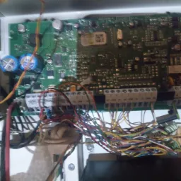 Otwarta obudowa centrali alarmowej z widoczną płytą główną i podłączonymi przewodami, widoczne kondensatory i złącza śrubowe.