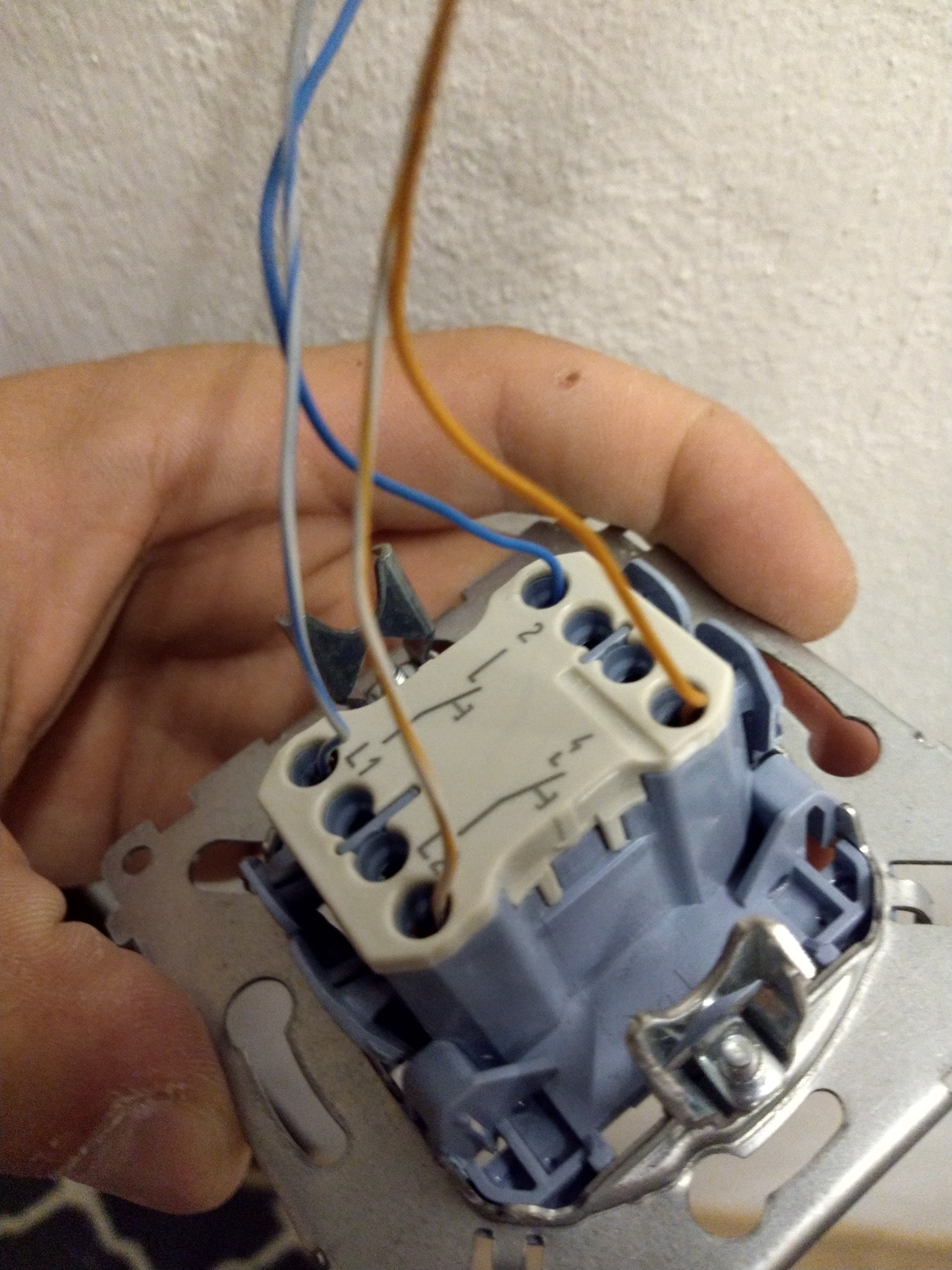 Dłoń trzyma podłączony, niebiesko-biały łącznik instalacyjny z wystającymi przewodami: niebieskim, szarym i pomarańczowym, na tle ściany.