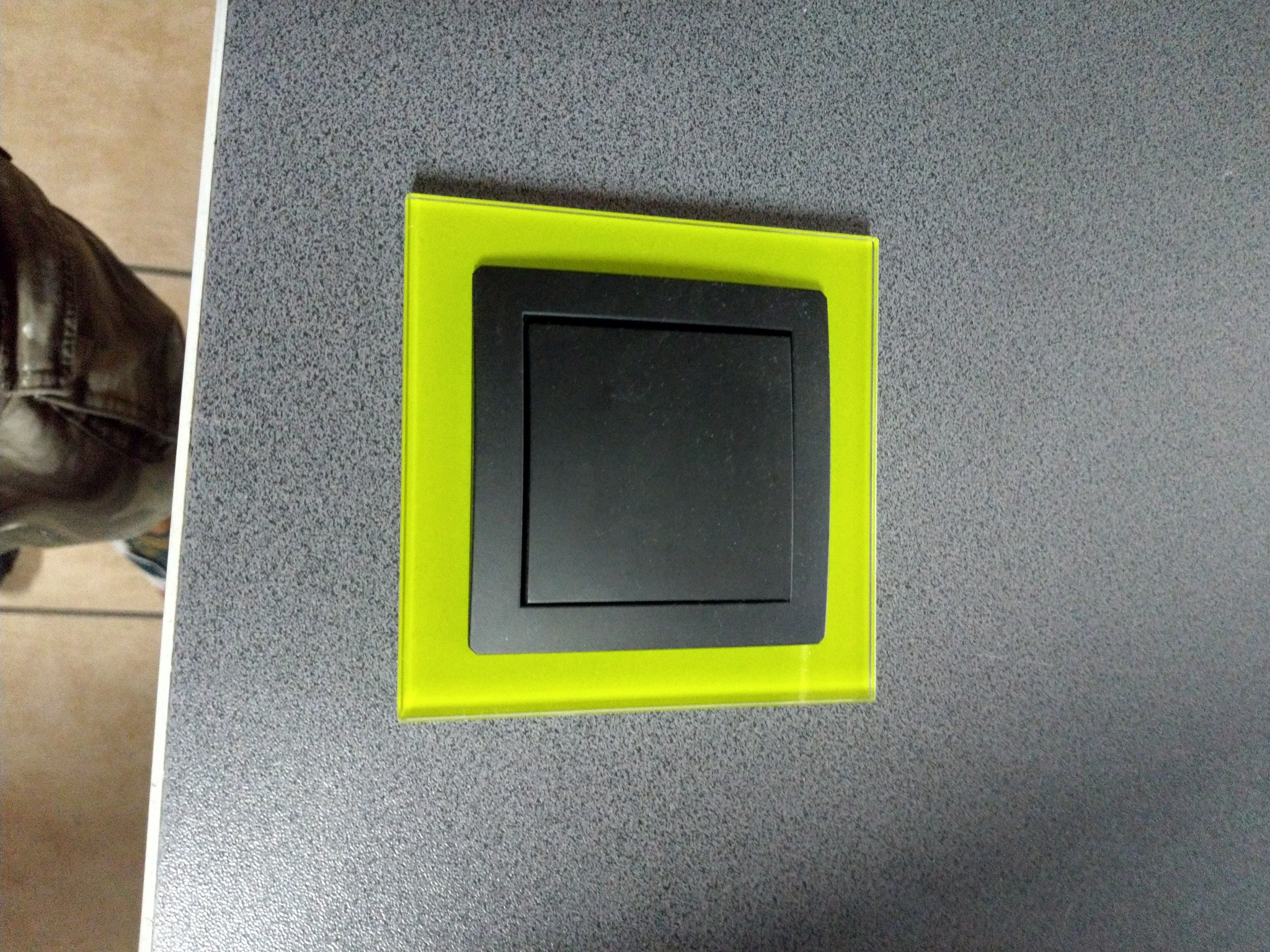 Czarny włącznik światła z podświetlaną, neonowo-zieloną ramką na szarej, teksturowanej ścianie.