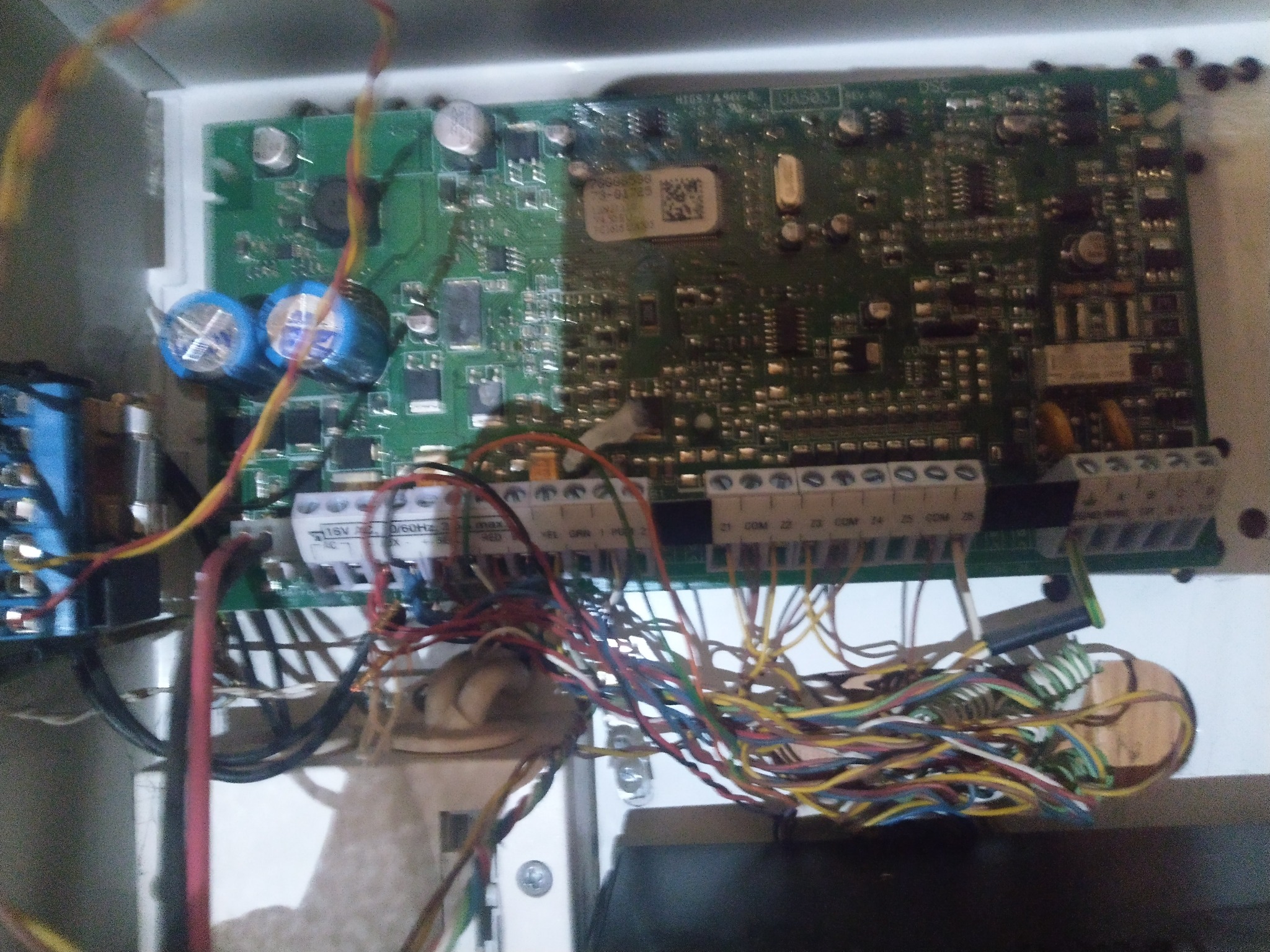 Otwarta obudowa centrali alarmowej z widoczną płytą główną i podłączonymi przewodami, widoczne kondensatory i złącza śrubowe.