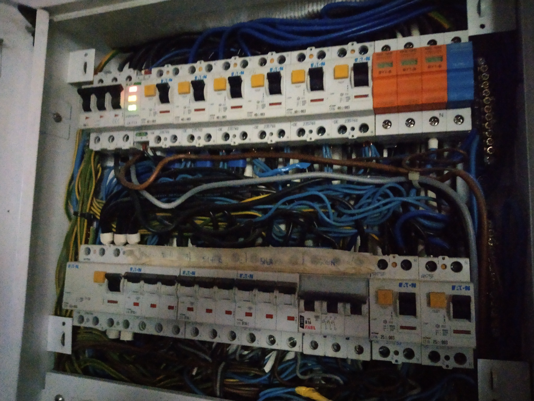 Otwarta skrzynka bezpiecznikowa z zainstalowanymi bezpiecznikami Eaton i Fael, widoczne liczne przewody w kolorach niebieskim, żółto-zielonym i czarnym.