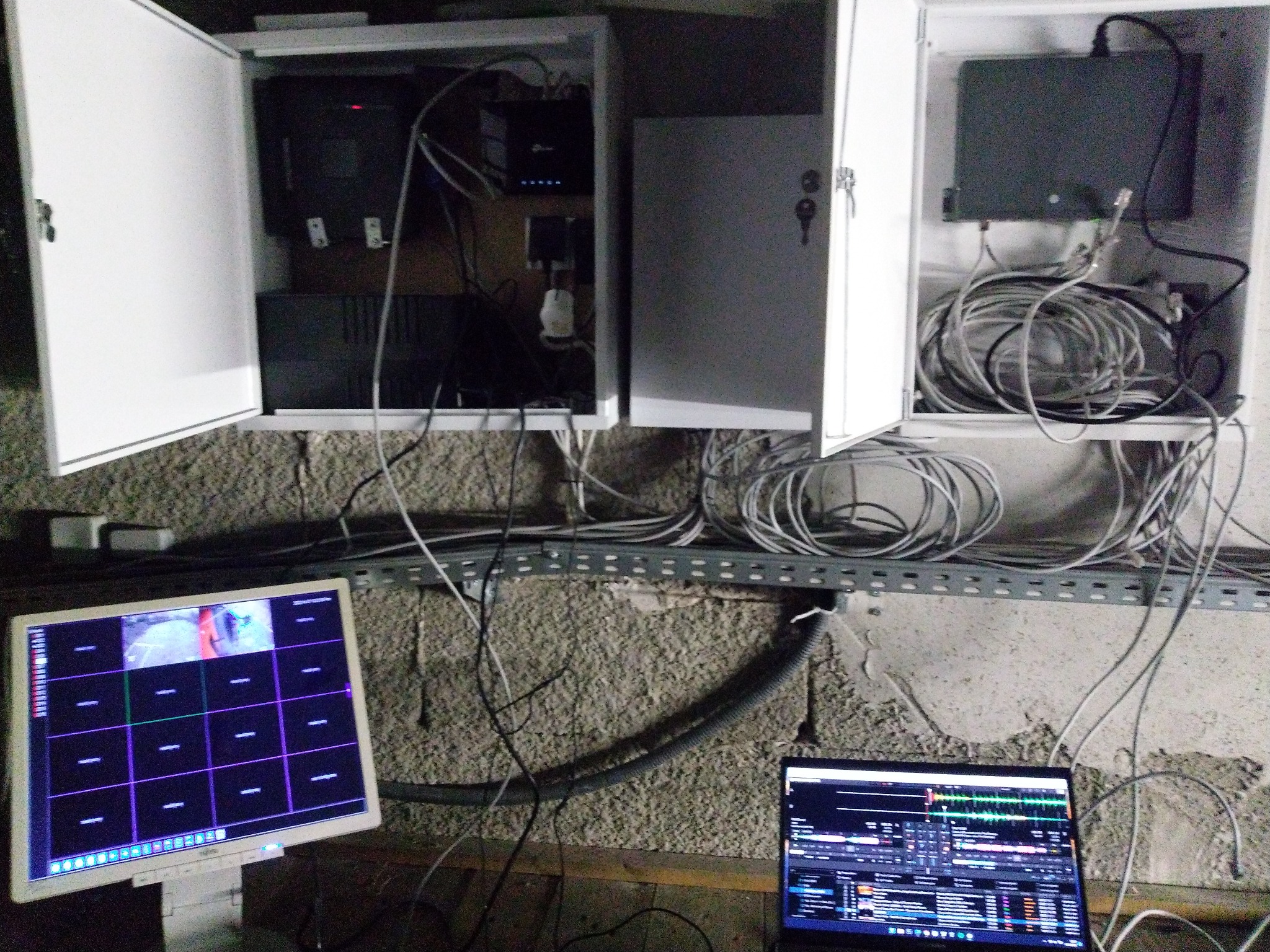 Dwa otwarte panele z zainstalowanymi urządzeniami elektronicznymi i plątaniną kabli, monitor i laptop wyświetlają obrazy z systemu kamer. Widoczna szyna montażowa na ścianie. Surowy stan...